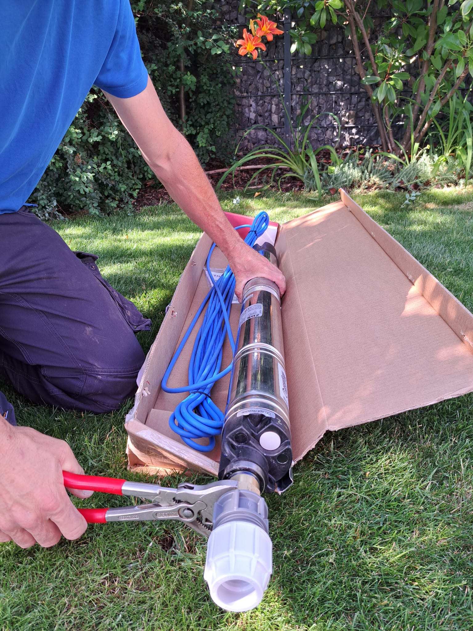Eine Person in einem blauen Hemd kniet im Gras und benutzt eine Zange, um eine Armatur an einer Metallpumpe mit einem blauen Kabel zu befestigen - perfekt für die Gartenbewässerung. Die Pumpe ist teilweise aus einem Pappkarton, im Hintergrund blüht ein Blumengarten.