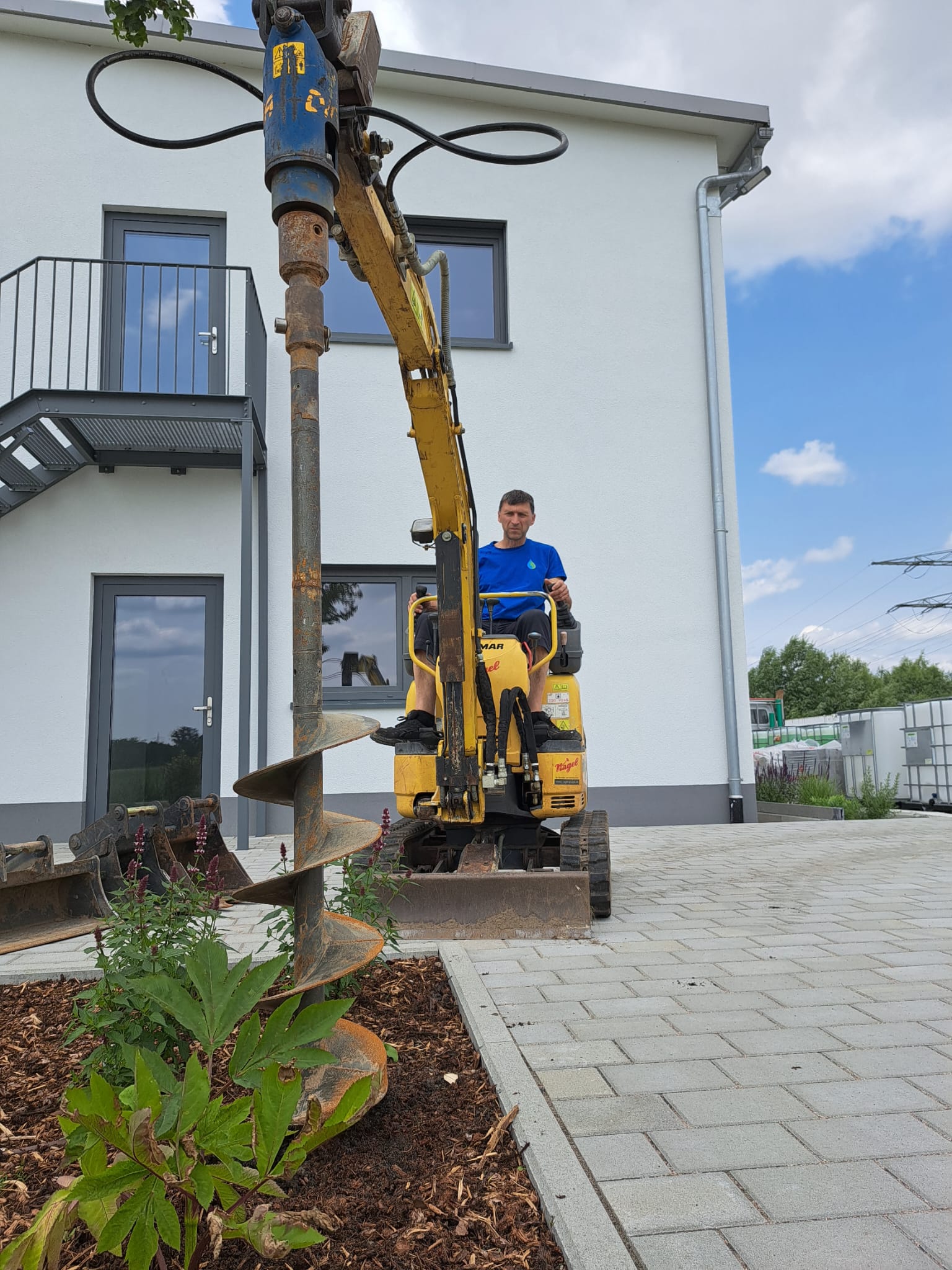 Ein Mann im blauen Hemd bedient einen gelben Minibagger mit einem Schneckenaufsatz, um den Boden für die Gartenbewässerung in der Nähe eines modernen weißen Gebäudes mit gepflastertem Gehweg und neu gepflanzten Sträuchern anzubohren.