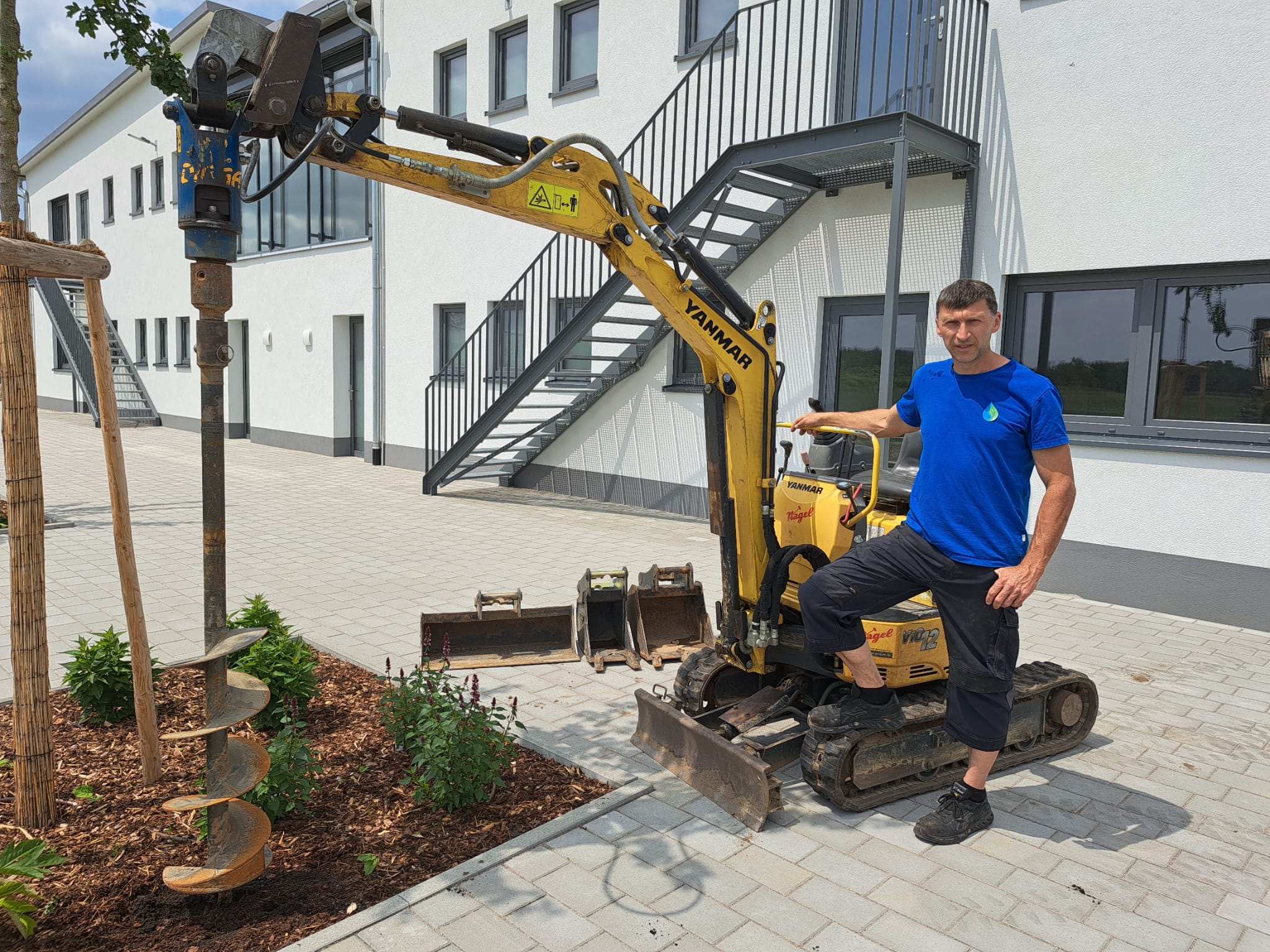 Ein Mann in einem blauen Hemd steht neben einem kleinen gelben Bagger mit einem Erdbohreraufsatz, der auf einer gepflasterten Fläche in der Nähe eines modernen weißen Gebäudes und eines angelegten Gartenbeetes abgestellt ist und möglicherweise die Installation einer Gartenbewässerung vorbereitet.